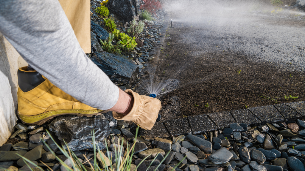 A person wearing work boots and gloves adjusts a broken sprinkler head in a garden